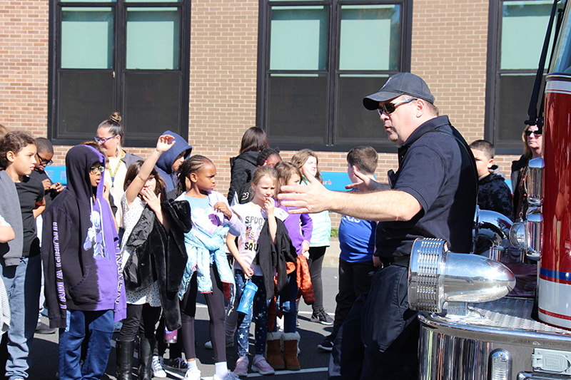 A man in a navy blue shirt, pants and baseball cap leans against a red fire engine and talks to a group of elementary students. One of the students is raising her hand.