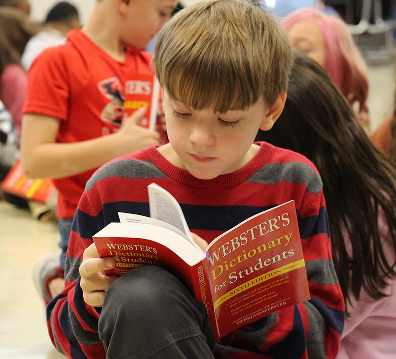 A boy with short sandy hair sits and looks through a dictionary. He is wearing a blue and red striped shirt.