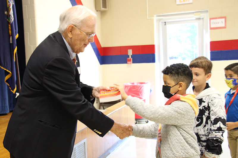 A man with white hair wearing a dark suit and tie hands a red dictionary to a young man and shakes his han. The boy is wearing a gray sweatshirt and has short dark hair. He is wearing a facemask.