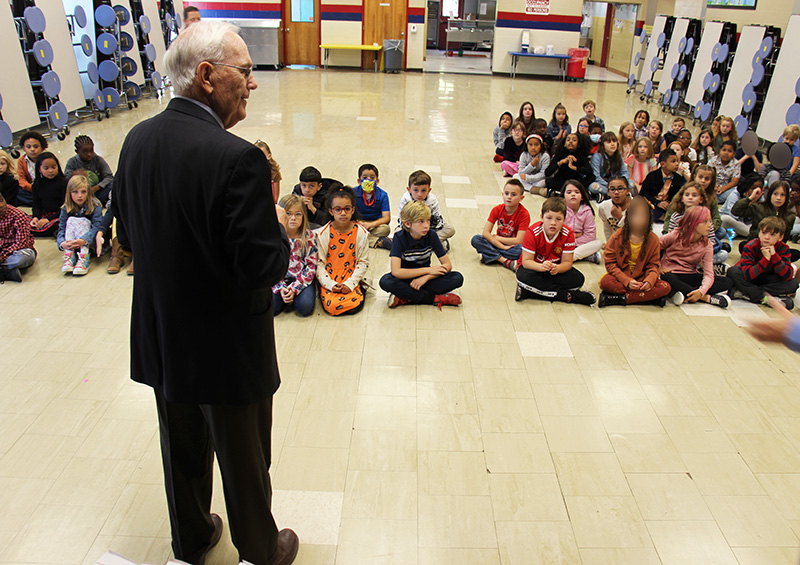 A man with white hair, wearing a dark suit stands in front of a group of third-grade kids talking to them.