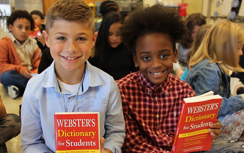 Two third-grade boys smile and hold up their dictionaries. The boy on the left has short sandy hair and is wearing a blue shirt. The boy on the right is wearing a red and black checkered shirt and has dark curly hair. Both are holding their red dictionaries up in front of them and are smiling.