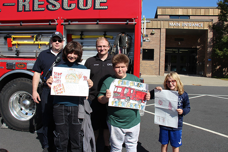 Two firefighters, dressed in navy blue shirts and pants, one with a blue baseball hat on, stand with three fifth-grade students who are holding fire safety posters they made. There is a fire truck in the background and a brown brick building and blue sky.