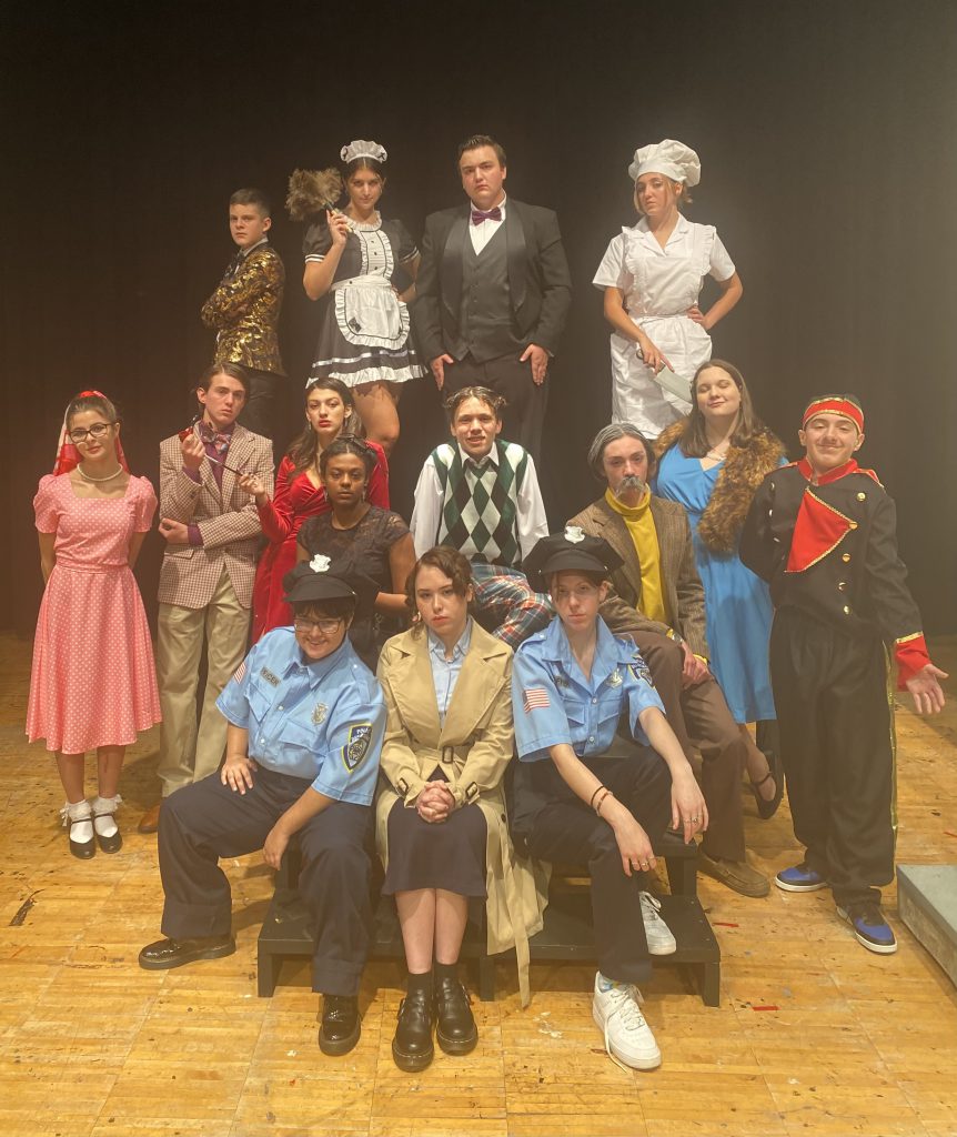 15 high school students dressed in costumes for the show Clue. Some are police officers, a maid, baker, doorman. 