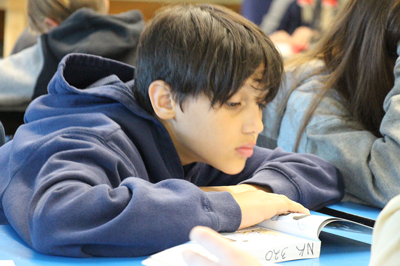 A sixth grade boy leans forward on his desk and reads a book. He has shaggy dark hair and is wearing a blue hooded sweatshirt.