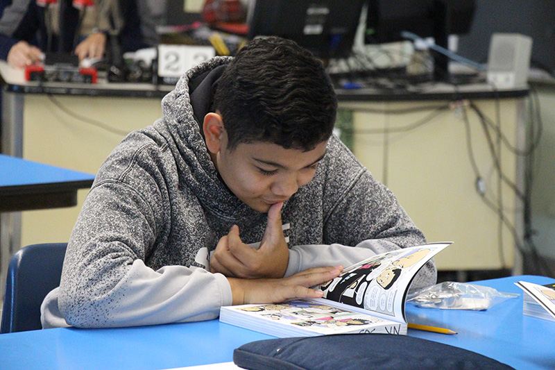 A sixth-grade boy with short dark hair and wearing a gray hoodie reads a book opened on his desk.