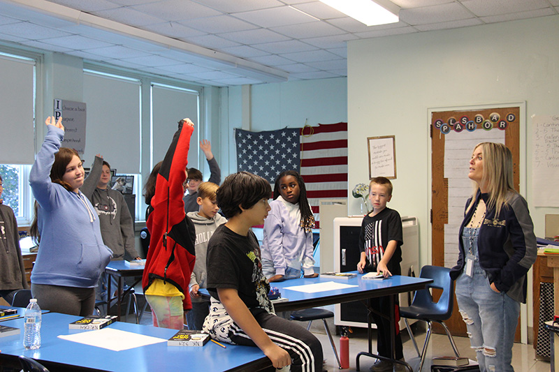 A classroom of sixth-grade students participate in a group activity. some are standing with their hands raised and others are sitting at their desks.
