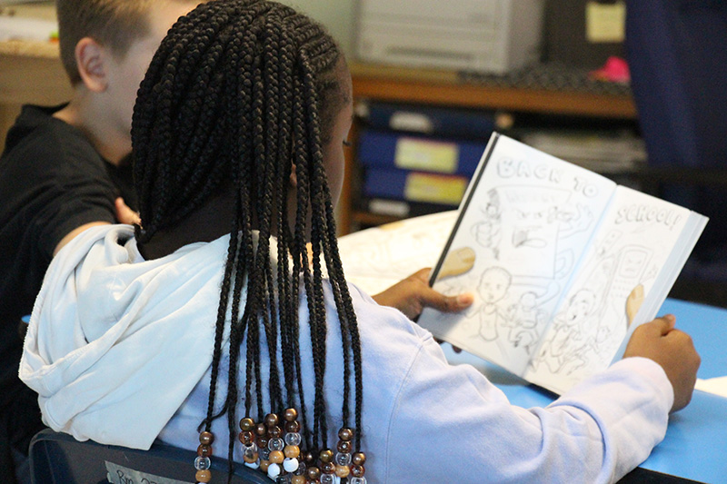 A sixth-grade girl with long dark braids  sits at her desk with a book open reading.
