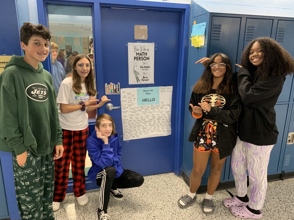 A group of five eighth grade students stand outside of a blue door that has a poster on it containing many different ways to say hello. The kids are pointing to the door and smiling.