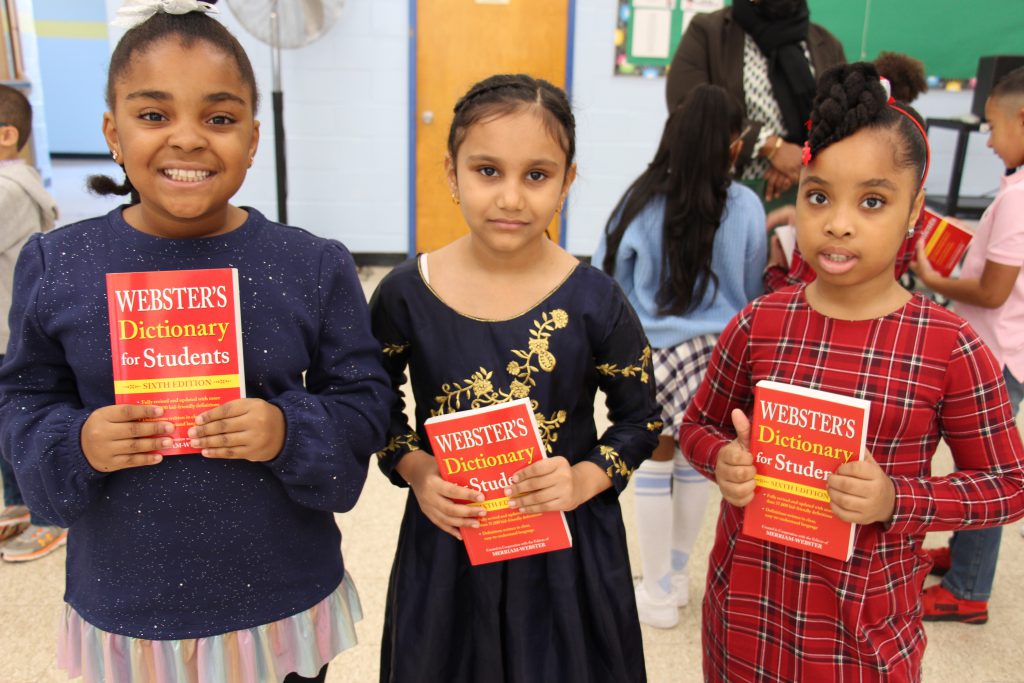 Three girls stand holding their red dictionaries. The girl on the left is smiling, her dar hair is pulled back and she is wearing a sparkly blue sweater. The girl in the center also has her dark hair pulled back and is wearing a blue dress with gold decoration on it. The girl on the right has her hair in a braid at th etop of her head, a red headband and is weraing a red plaid dress.