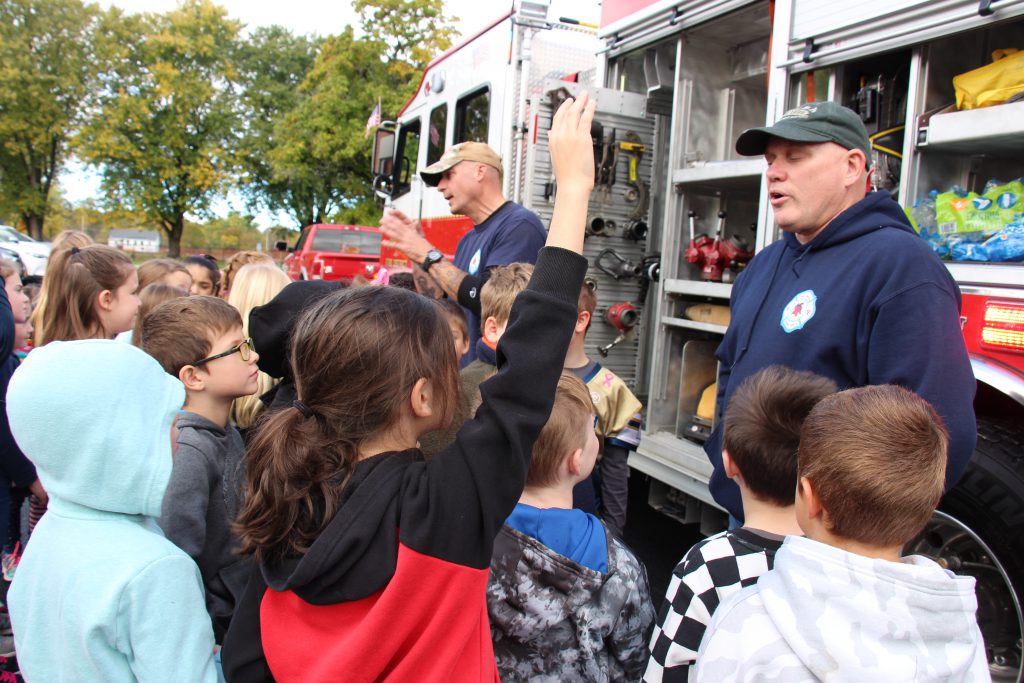 A red fire truck says Pine Bush 228 in the background. There ar etwo men wearing navy blue shirts standing talking to a group of elementary students. One of the students has her hand up.