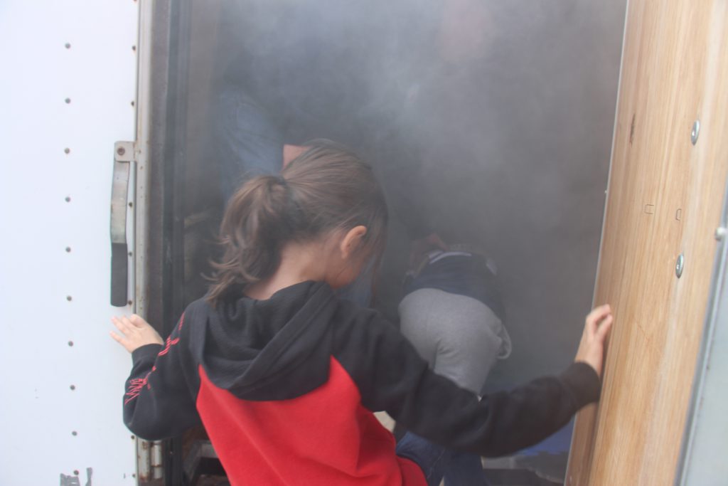 An elementary age student wearing a black and red hoodie goes into a room that is simulated smoky.