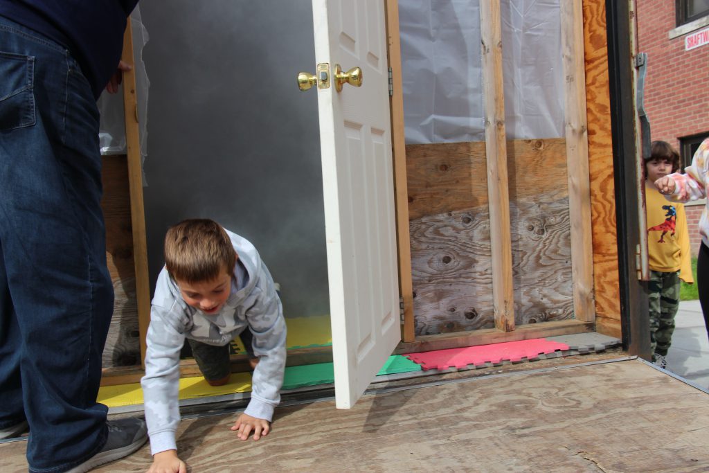 A boy with short sandy hair wearing a gray sweatshirt crawls out of a simulated smoky room.