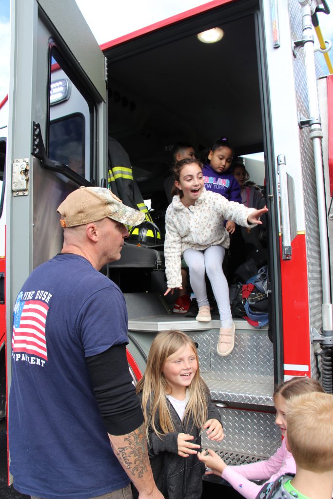 An elementary-aged girl puts her hand out to grab a handle as she leaves a fire engine. There are kids in front of her and behind her. A man wearing a blue shirt with an american flag on the back stands watching her.