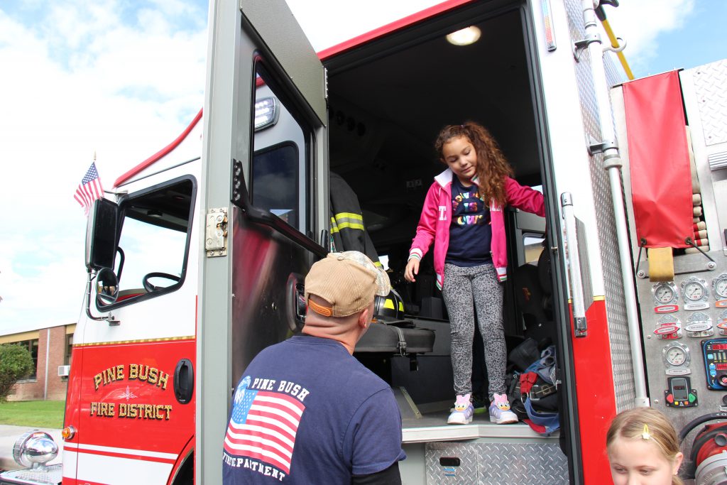 An elementary-aged student with long brown hair, wearing a pink jacket, stands at the top of the stairs of a fire engine as she starts to leave. A man wearing a blue shirt with an american flag on it stands ready to help her.
