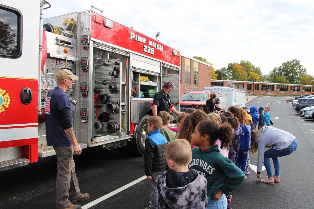 Two firefighters wearing navy blue shirts stand in front of a red fire truck that says Pine Bush. There is a class of elementary aged kids listening to them talk.