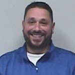 A man with short dark hair, close beard and mustache, wearing a blue shirt with a white shirt under it, smiles.