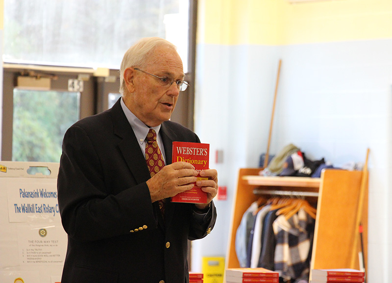 A man wearing a dark suit and red tie holds a red dictionary up as he talks to a group of children.