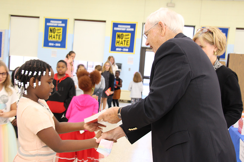 A man with white hair, wearing a dark suit shakes hands with a third-grade girl and hands her a red dictionary. The girl is dressed in white and has white beads in her braided hair.