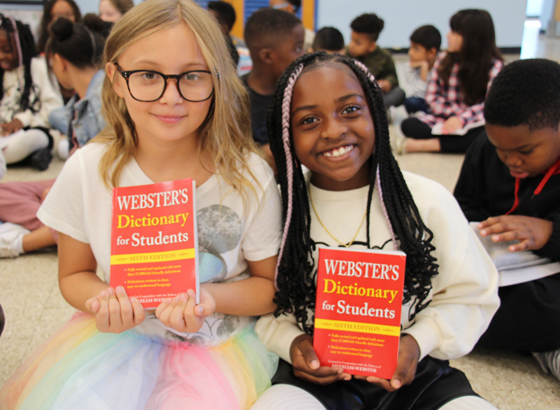 Two third-grade girls hold red dictionaries. They are sitting on the floor among other students. They are smiling. The girl on the left has shoulder-length blonde hair and is wearing glasses and a multi-color skirt. The girl on the right has a white sweater on and her long dark braids have a bit of purple in them.