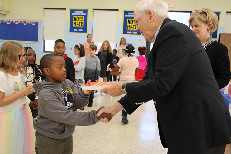 A third-grade boy in a gray sweatshirt shakes hands with a man with white hair wearing glasses and a dark suit. The man is giving him a red dictionary.