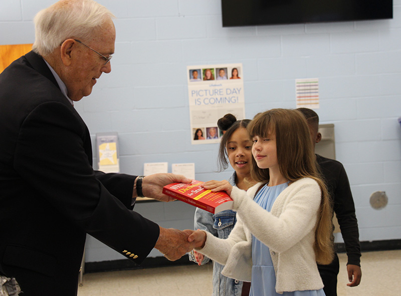 A girl with a light blue shirt, white sweater and long brown hair shakes hands with a man with white hair wearing glasses and a dark suit. He is handing her a red dictionary.