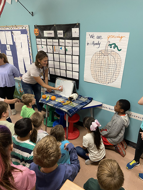 A woman stands in front of a class of fourth-grade students. There is a table next to her with fruit, carrots and celery on it, as well as a multi-colored guinea pig. There is a white board with all of the foods listed on it. 