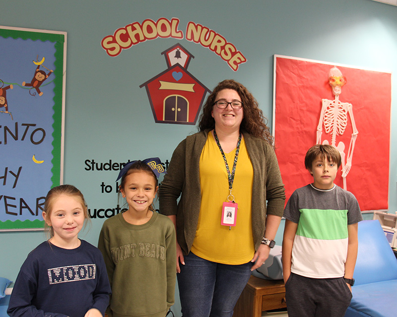A woman with long dark hair and glasses, waring a brown sweater and gold shirt smiles under a sign that says School Nurse with a little red schoolhouse by it. There are three children standing with her smiling. The girl on the left has her long hair pulled back into a ponytail and is weark a blue shirt that says mood. The girl next to her has her dark hair pulled back and is wearing a green shirt. Then is the woman. A boy on the right has short dark hair and is wearing a short-sleeve shirt that is green and white. 