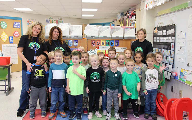 Three teachers all wearing black t shirts with green rainbows on them stand with a class of kindergarten students, about 18 of them. The kids are wearing shades of green.