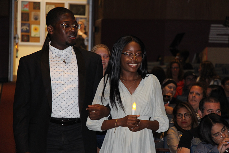 A young man is on the left, wearing a white shirt and black jacket. On the right is a young woman holding a battery-operated candle. She has long dark hair and is wearing a gray dress. She is smiling broadly.