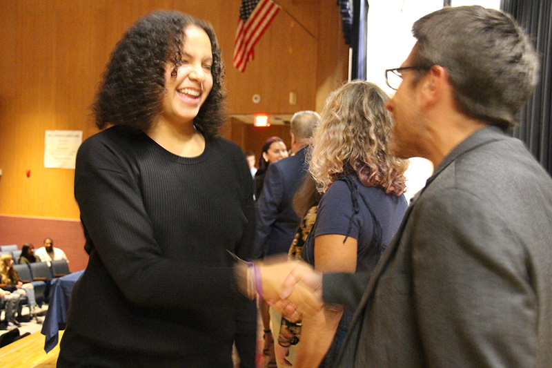 A young woman with shoulder-length curly black hair smiles and shakes the hand of a man wearing a gray jacket and glasses. The young woman is wearing a black dress. There are people behind them also shaking hands.