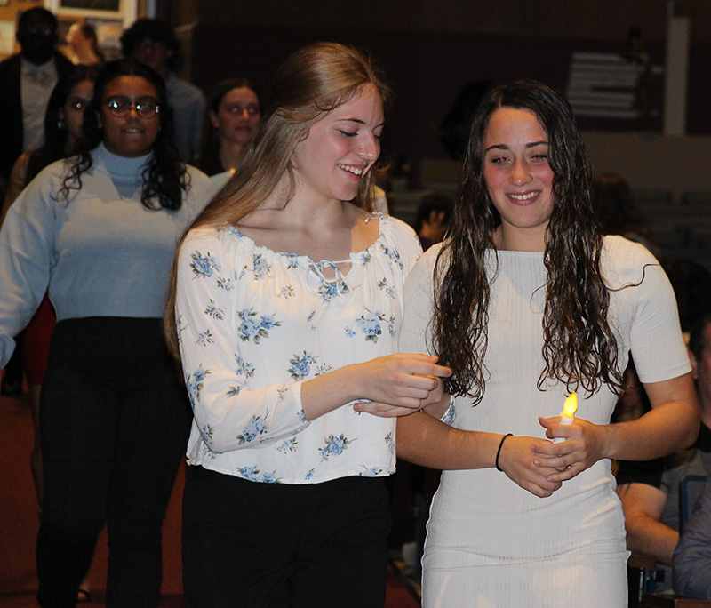 Two high school young women walk down an aisle,hooking arms together. The girl on th eleft has long blonde hair and is wearing a white printed shirt and black pants. The girl on the righ thas long dark hair and is wearing a gray dress. She is holding a battery operated candle.+++++++++