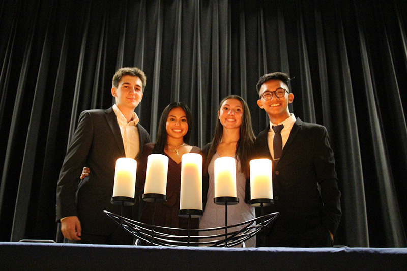 Four high school students stand behind five large pillar candles. A young man on the left is wearing a jacket and dress shirt. He has short dark hair. The young woman next to him has long dark hair and is smiling. The young woman next to her has long dark hair and is wearing a light colored dress. She is smiling. The young man on the right side is wearing a dark suit and tie. He has glasses and dark hair. There is an auditorium curtain behind them.