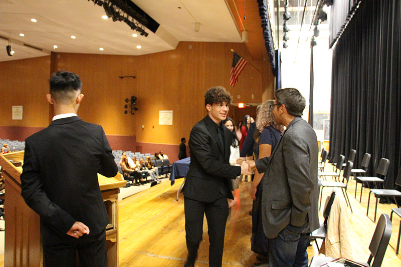 A young man shakes the hand of a man on a stage. The young man is wearing a dark suit and has dark hair. The man is wearing a suite and glasses. 