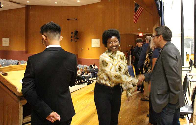 A young woman shakes the hand of a man on a stage. The young woman is wearing dark pants and a lighter printed shirt. She has short dark hair and is smiling.