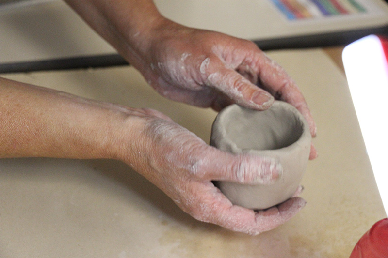 A pair of hands mold gray clay into a small pot. The hands have clay on the fingers.