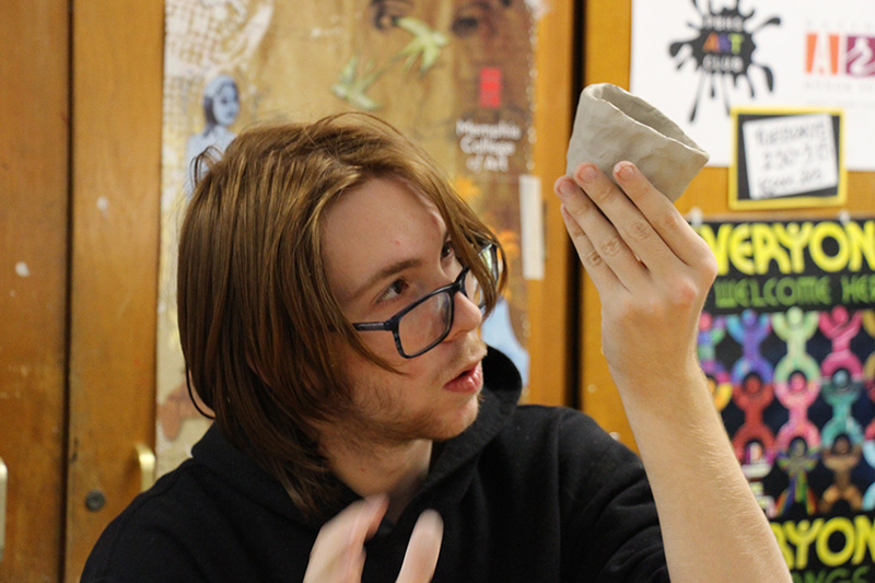 A high school student with chin-length reddish hair and glasses, holds up a clay pinch pot so he can see the bottom as he is making it.