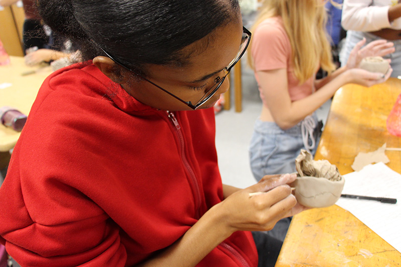 A young woman in a red sweatshirt, with dark hair pulled back and glasses, molds her clay into a pot.