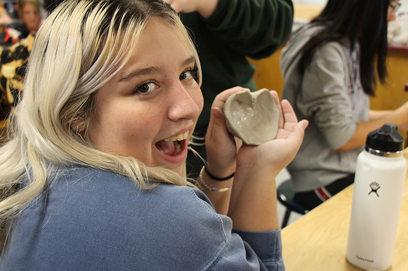 A young woman with mid-length blonde hair smiles and holds up a small clay pot she shaped into a heart.
