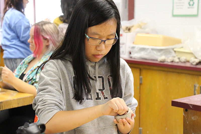 A young woman with long black hair, wearing a gray sweatshirt, molds her clay into a small pot.