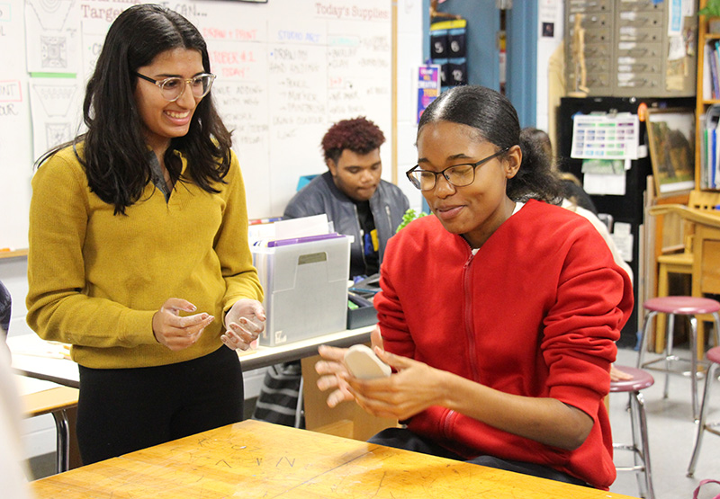 Two girls are at a table. The girl on the left has long dark hair and is wearing glasses and a mustard colored shirt. She is smiling and watching the girl on the right, who is sitting, as she molds clay in her hands.