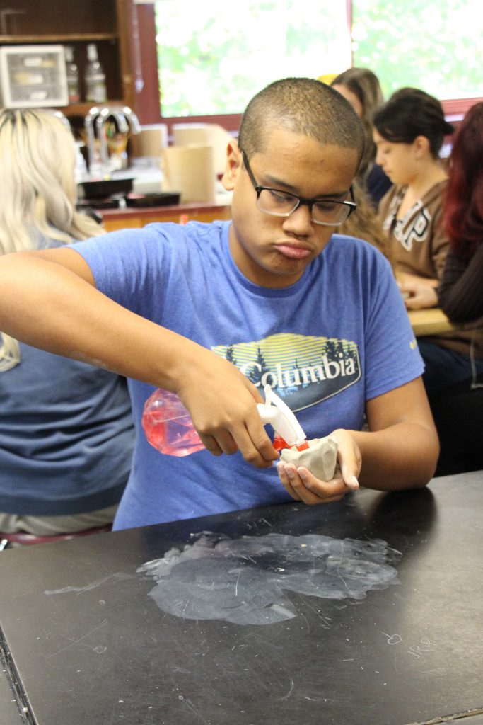 A boy with short dark hair, glasses and wearing a blue tshirt squirts a spray bottle of water into a small clay pot.