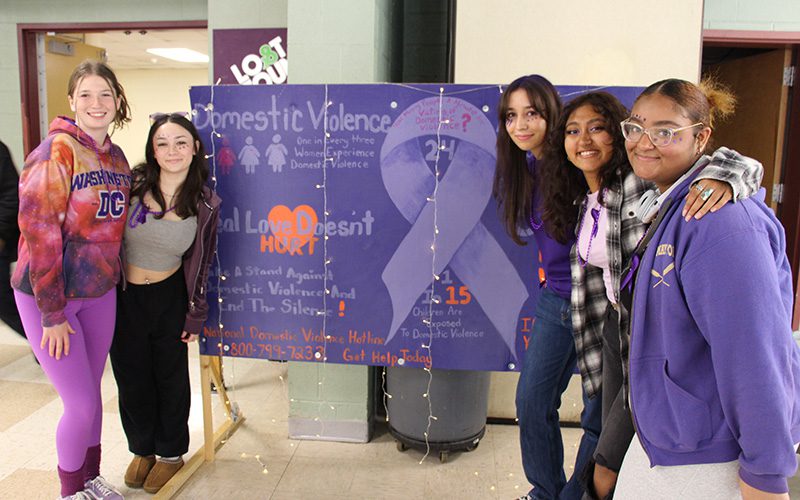 A group of five girls - two on the left and three on the right - flank a large purple sign about domestic violence.