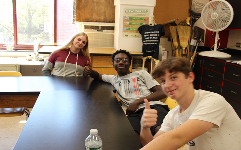 Three high school students sit in a classroom at an L shaped table. All are smiling. Two boys on the right are giving thumbs up. The girl on the left has long blonde hair.