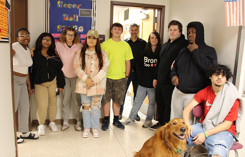 A group of nine high school kids and one man stand in a semi-circle in front of a bulletin board that says STARS Honor Roll. There is a reddish long-haired dog sitting in the front.