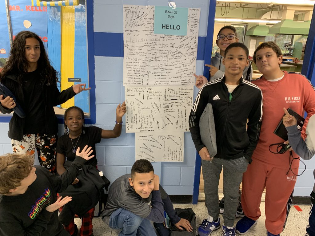 A group of seven seventh-grade students stand in a hallway and point to a very large list of words on a long poster. These are all ways to say hello in different languages. 