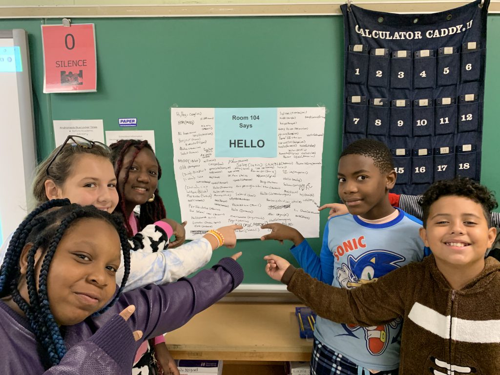 Five sixth grade students point to a poster that is on a blackboard. On the poster are ways to say hello in many languages.