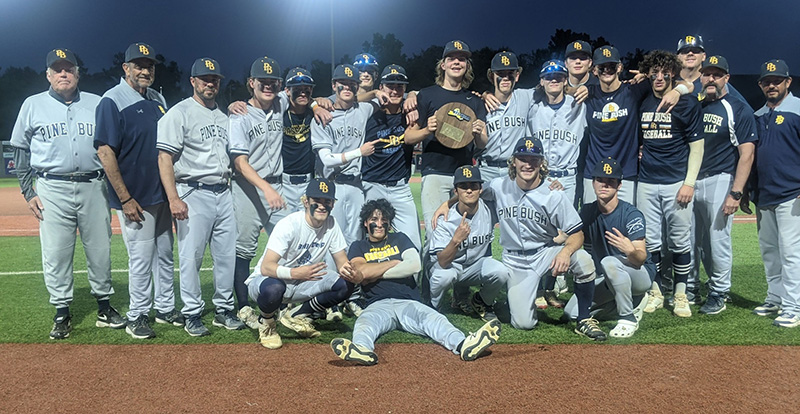 A group of 22 high school boys and coaches, all dressed in white baseball uniforms with blue caps. There are five kneeling in front and the rest standing behind. The boy in the center is holding a round plaque.