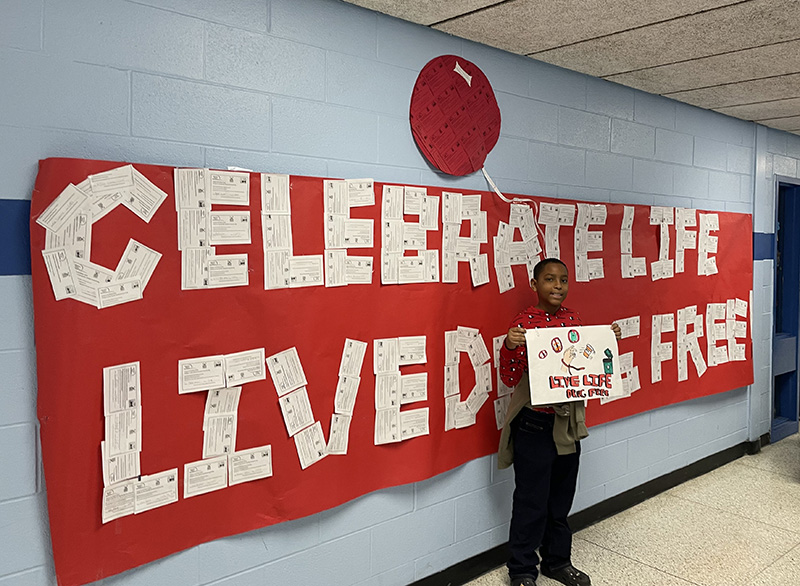 A middle school student holds up a poster he made that says live life drug free. Behind him is a large red poster with white cards arranged so it says Celebrate Life Live Drug Free.