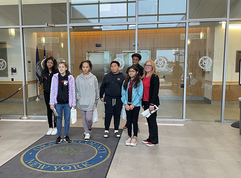 A group of six middle school students and an adult stand together with a glass door behind them and a rug below that says Orange County NY