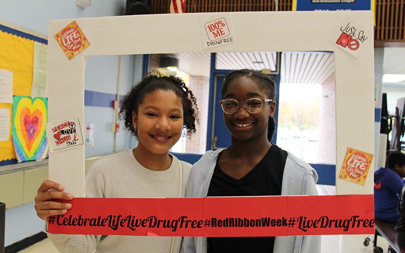 Two middle school girls smile and hold up a picture frame around them that says Celebrate life live drug free.